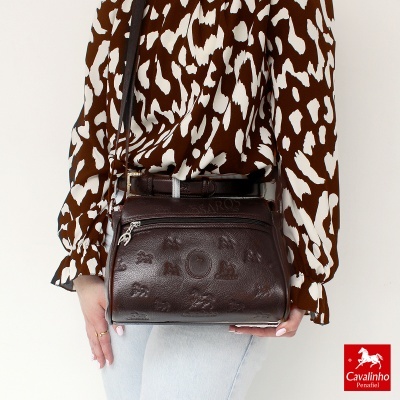 Bolsa de pele castanha com padrão em relevo de cavalos e texto Cavalinho, com alça ajustável, usada por pessoa com blusa castanha e branca