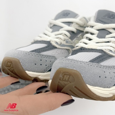 Ténis New Balance cinza e branco com sola castanha e atacadores brancos.