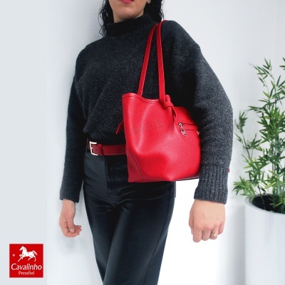 Mala vermelha em pele com zíper prateado e alças duplas, usada por mulher com camisola cinza e calças pretas.
