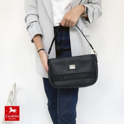 Mala preta Cavalinho com alça em mão de pessoa com casaco cinza e jeans azul