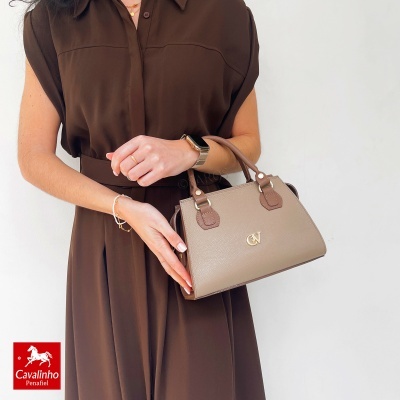 Mala de mão bege com detalhes castanhos e logotipo dourado segurada por mulher com vestido castanho