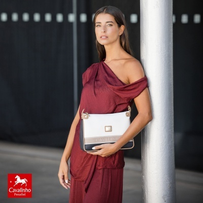 Mulher com vestido vinho segura mala Cavalinho branca, bege e preta e está apoiada numa coluna branca.