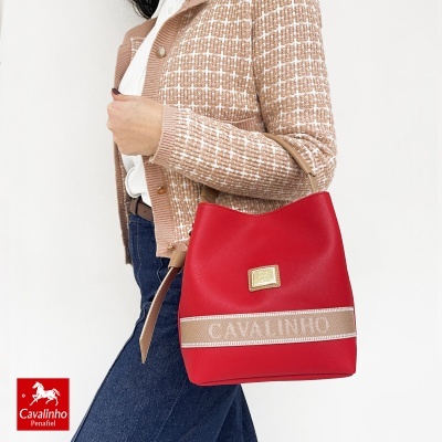 Mala vermelha Cavalinho de material sintético com detalhes bege e dourados, segurada por mulher com casaco bege xadrez e jeans.