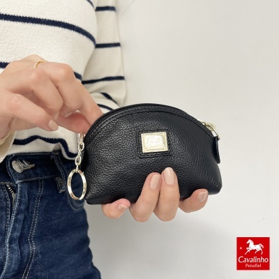 Carteira preta em pele texturada com fecho e argola metálica, segurada por mão vestindo camisola de riscas azuis e jeans.