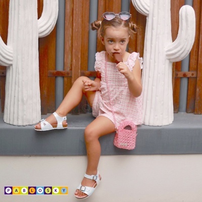 Criança sentada com vestido rosa, sandálias brancas e bolsa rosa junto a cactos brancos