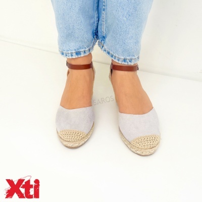 Sapatos espadrilha cinza com tiras castanhas no tornozelo e biqueira em ráfia numa superfície branca