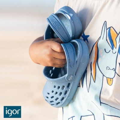 Calçado infantil azul claro IGOR em borracha com alça e furos, sendo segurado por criança com t-shirt de unicórnio.