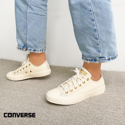 Ténis Converse All Star creme em pele, com atacadores dourados, calças de ganga azul claro e carpete cinzenta