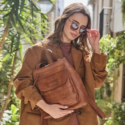 Mochila castanha em couro com fecho zip segura por mulher com casaco castanho claro e sunglasses em ambiente exterior