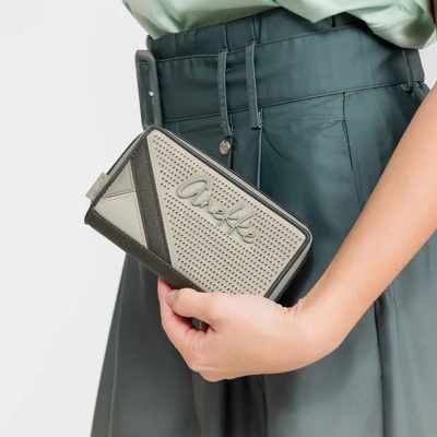 Carteira cinza com padrão perfurado e nome Anekke, segurada por mão, com calças verdes.