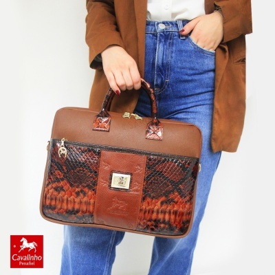 Mala castanha com textura de pele de cobra e alças curtas, mostrada por mulher vestindo casaco castanho e jeans azuis.