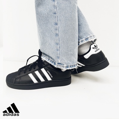 Tênis Adidas Superstar preto e branco com riscas brancas e logo Adidas, usado com calças de ganga claras.