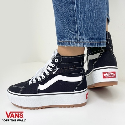 Ténis Vans pretos de cano alto com risca branca e sola branca, usados por pessoa com jeans azuis.