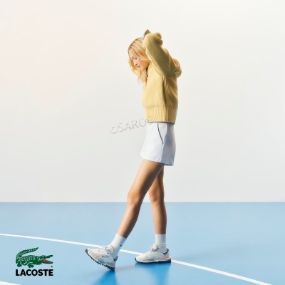 Mulher vestindo camisola de malha amarelo mostarda, saia branca e ténis brancos em piso azul com logo Lacoste.