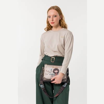 Mulher com camisa bege e calças verdes segurando bolsa bege estampada Anekke.