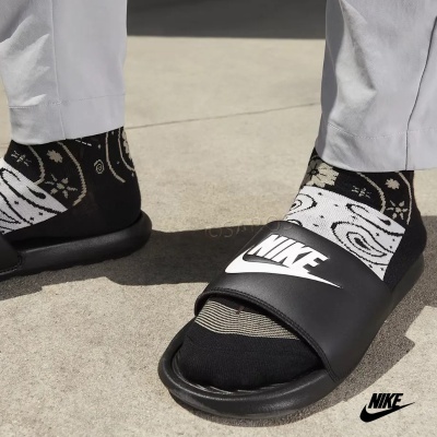 Chinelos Nike pretos com meias pretas e brancas e calças cinza claro num pavimento exterior.