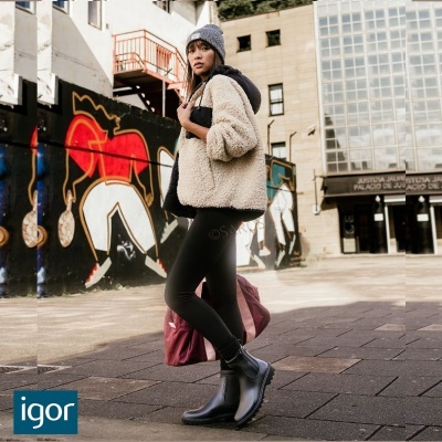 Mulher vestindo casaco peludo bege com capuz preto, leggings pretas, botas pretas e gorro cinzento em rua com arte urbana.
