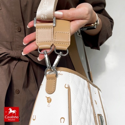Mala branca com padrão bege, alça larga bege e fechos metálicos, segurada por mão com unhas brancas