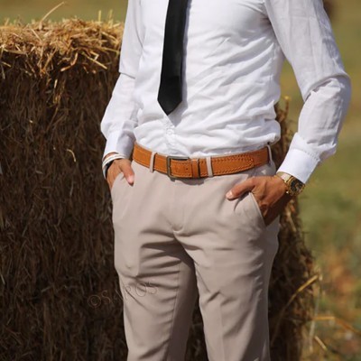 Homem com camisa branca, gravata preta, cinto castanho e calças bege junto a palha