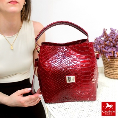 Mala vermelha texturada com alça e fecho dourado, apresentada em mesa branca ao lado de mulher e vaso de flores roxas.