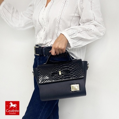 Mala preta de mão com textura de pele de cobra e fecho dourado, usada por pessoa com camisa branca e calças azuis