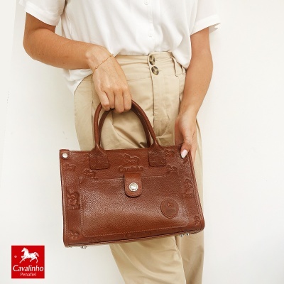 Mala de mão castanha em pele com detalhe de cavalos, segurada por pessoa com roupa clara