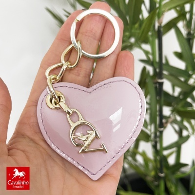 Chaveiro coração rosa verniz com letras douradas e corrente, mão segurando, fundo folhas verdes