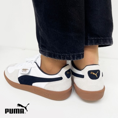 Ténis Puma brancos com detalhes castanhos e azul escuro, pessoa de calças pretas, fundo branco