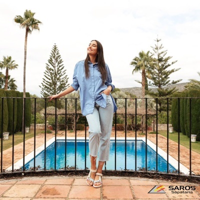 Mulher com camisa azul clara, calças cinza e sandálias brancas junto a piscina e cerca preta