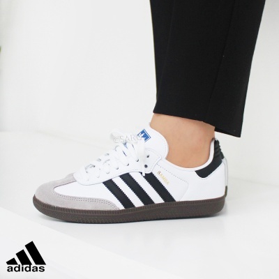 Ténis Adidas brancos com riscas pretas e sola castanha usados num pé com calças pretas