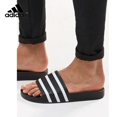 Chinelos Adidas pretos com riscas brancas usados por pessoa com calças pretas.
