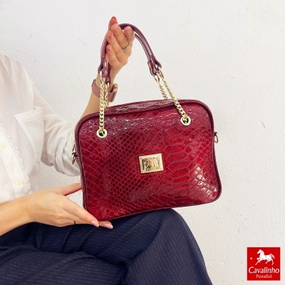 Mala de mão vermelha com textura de pele de cobra e alças douradas, segurada por uma pessoa.