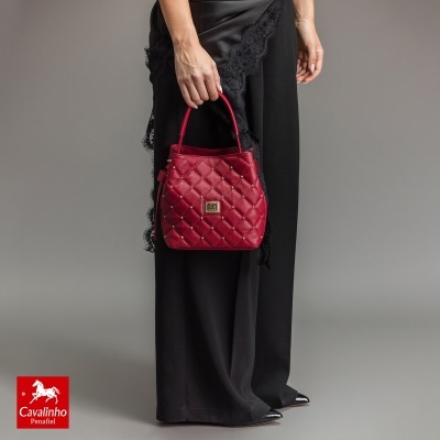 Bolsa de mão vermelha com padrão matelassê e rebites dourados na mão de uma mulher com calças pretas rendadas.