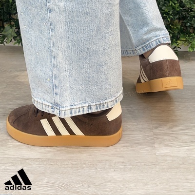 Ténis Adidas castanhos com riscas brancas em uso com calças de ganga claras num interior com plantas verdes.