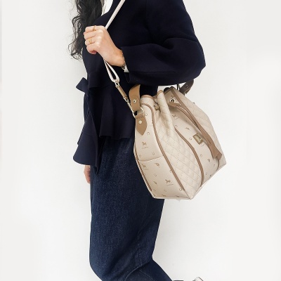 Mala de ombro bege com padrão acolchoado e detalhes castanhos, usada por pessoa com casaco preto e calças azuis.