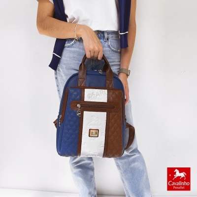 Mochila Cavalinho azul, castanho e branco com padrão matelassé e logótipo, segurada por pessoa com roupa casual.