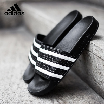 Chinelos Adidas pretos com riscas brancas em piso de pedra cinzenta.