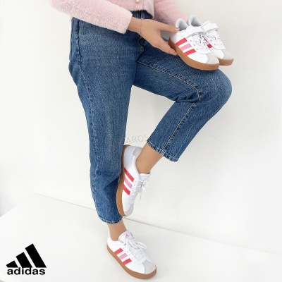 Sapatilhas Adidas brancas com riscas vermelhas em uso e em mãos, vestidas com calças de ganga azuis.