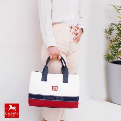 Mala de mão branca, azul e vermelha com alças pretas, com texto CAVALINHO, segurada por pessoa vestindo camisa branca e calças claras.