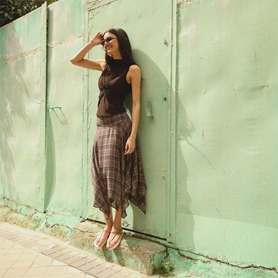 Mulher com blusa preta sem mangas e saia xadrez posando junto a parede verde clara.