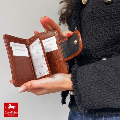 Carteira castanha de pele aberta com fecho, segura por mãos femininas vestindo casaco preto e jeans.