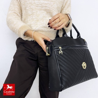 Mala preta com padrão matelassé e detalhes dourados, segurada por mulher com casaco bege e calças pretas