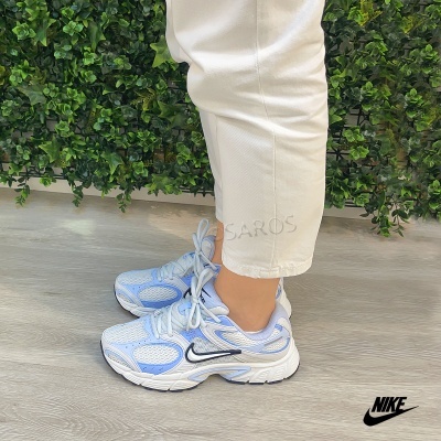 Ténis Nike brancos e azuis com detalhe preto usados com calças brancas e fundo verde de plantas