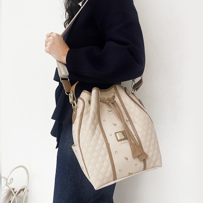 Mala tipo saco bege com padrão matelassé e símbolos, detalhes em castanho claro, alça larga bege e placa dourada, usada por uma pessoa.