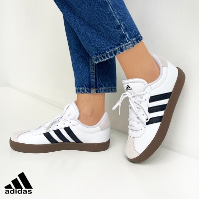 Tênis Adidas branco com riscas pretas e sola castanha usados com jeans azuis