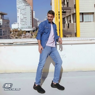 Homem com camisa de ganga azul, t-shirt rosa clara, jeans azul claro e sapatos pretos Skechers a posar em ambiente urbano