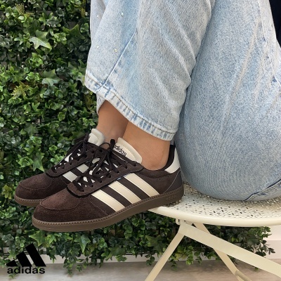 Ténis Adidas castanhos de camurça com riscas brancas e calças de ganga claros sentado numa cadeira branca com fundo de plantas verdes.