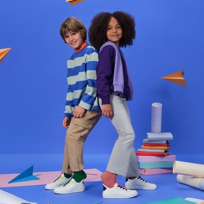 Duas crianças vestidas casual com camisola e ténis brancos sobre fundo azul com aviões de papel.