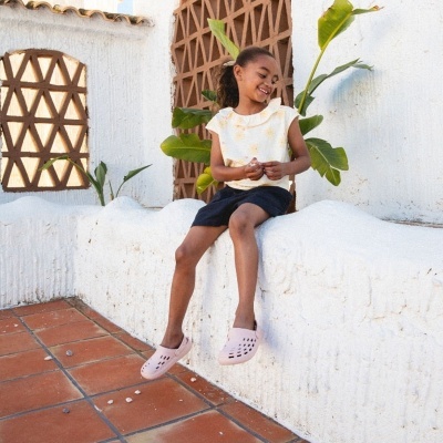 Criança sentada numa parede branca com plantas ao fundo, vestindo camiseta branca com flores amarelas, calções pretos e sapatos rosa claro.