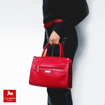 Mala de mão vermelha de pele com fecho numa mão, pessoa com roupa preta e cinto vermelho